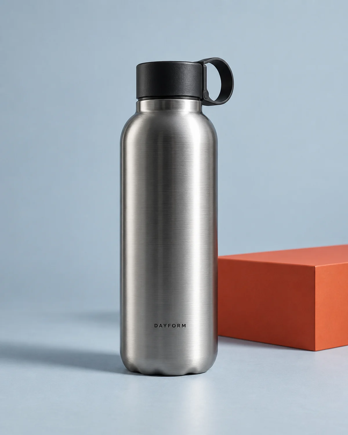 Fictional DAYFORM brushed-steel bottle with a black carry lid on a sculptural orange studio plinth